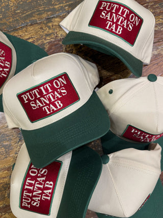 Pile of trucker hats including a cream and forest green hat embroidered Put It On Santa's Tab