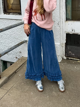 Woman wearing the Lane denim blue corduroy wide leg pants with a layered ruffle hem and a pink deer sweater