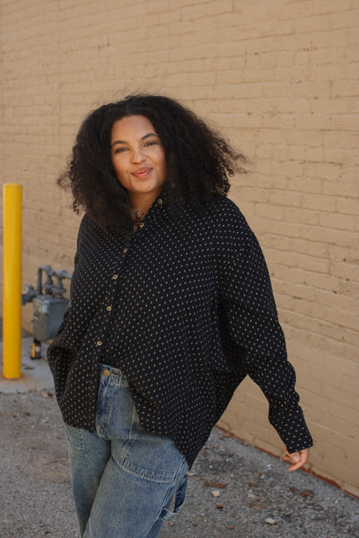 Woman wearing a black patterned shirt and jeans standing against a beige wall.