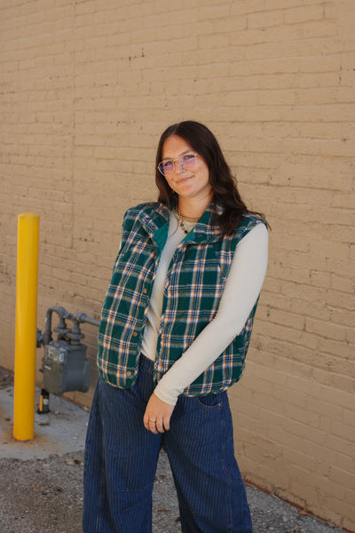 Person wearing a plaid vest and blue pants standing against a beige brick wall.