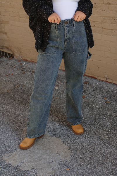Person wearing blue jeans and a dark jacket standing on a concrete surface with a beige wall in the background.
