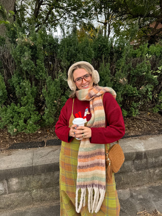 Woman wearing a brushed stripe fringe scarf in cream, orange and sage over a red sweater, with earmuffs and a plaid overall dress