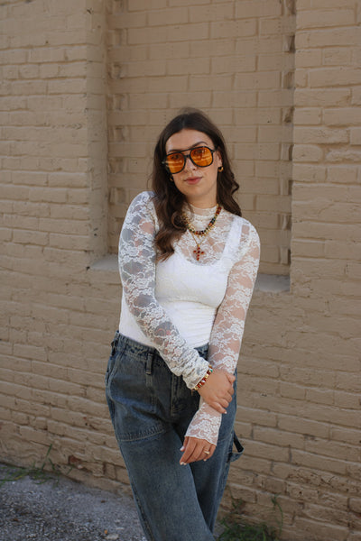 Woman wearing a ivory lace top and blue jeans against a beige brick wall.