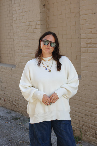 Person wearing a white sweater and sunglasses standing against a brick wall.