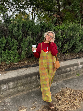 Woman wearing the Reese washed olive plaid oversized cotton jumpsuit with adjustable straps over a red sweater