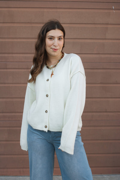 Woman wearing a cream cardigan and blue jeans standing against a brown wall.