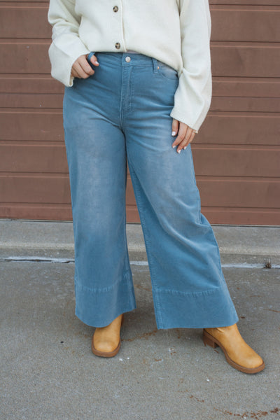 Girl wearing blue corduroy wide leg jeans and a cream cardigan against a brown wall.