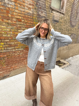 Woman wearing the Arlo heather gray winter knit cardigan with a cream snowflake yoke and wooden buttons with brown striped wide leg pants