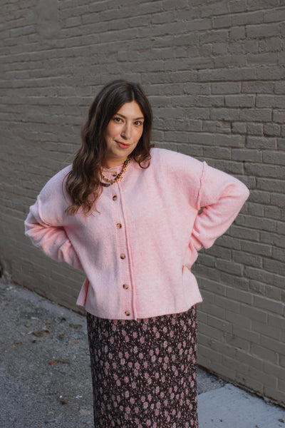 Woman wearing a pink cardigan and floral skirt against a gray brick wall.
