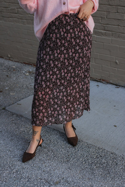 Person wearing a pink shirt and floral skirt standing on a sidewalk.