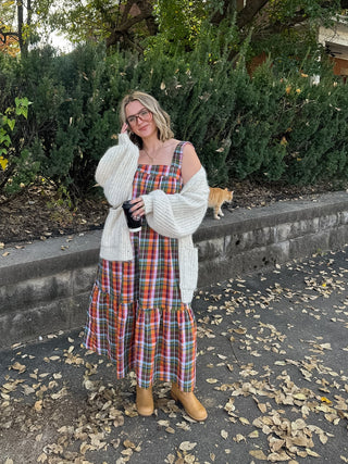 Woman wearing a rainbow plaid tiered maxi dress with a cream chunky cardigan and tan boots, holding a coffee
