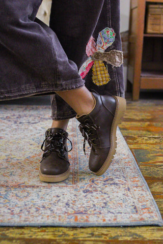 Woman wearing Corkys Scout Campfire walnut distressed faux leather lace up ankle boots with a lug sole and brown corduroy pants