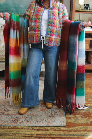 Woman holding two oversized brushed plaid fringe scarves in rainbow and navy multi colors, wearing a quilted plaid jacket, white tee and wide leg jeans