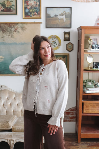 Woman wearing the Remy ivory knit tie front cardigan with embroidered holiday bows, ornaments and holly and brown corduroy pants