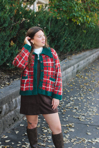 Woman wearing a red and green chunky plaid cardigan with a pointed collar over a white turtleneck and brown suede jumper with tall brown boots