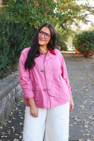 Woman wearing a pink oversized barn jacket with a red corduroy collar and metal clasp closures over cream wide leg pants