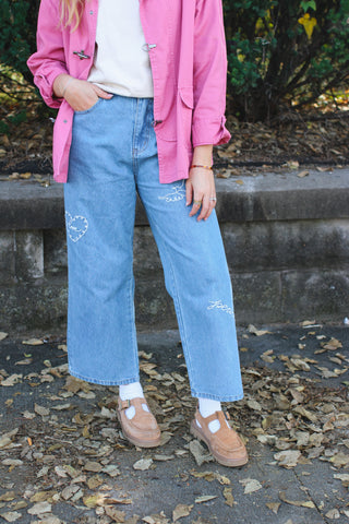 Woman wearing light wash wide leg jeans with white embroidered heart, dove and peace script, a pink jacket and tan suede T-strap shoes with white socks