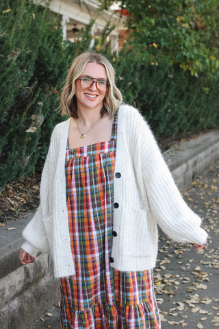 Woman wearing an ivory oversized knit cardigan with black buttons and pockets over a plaid maxi dress