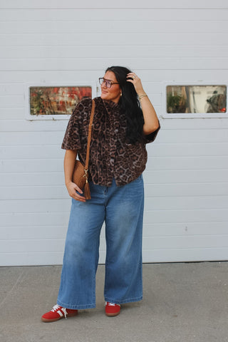 Woman wearing a faux fur leopard print capelet with a funnel neck and pockets over wide leg jeans and red sneakers