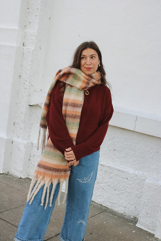 Woman wearing a burgundy knit pullover sweater with a colorful striped fringe scarf and embroidered wide leg jeans