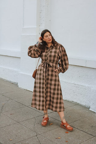 Woman wearing an olive and blush pink plaid midi shirtdress with puff sleeves and a tie waist with tan wedge shoes
