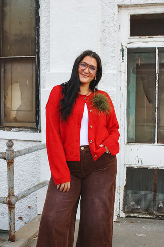 Woman wearing a red button front knit cardigan with an olive green feather brooch over a white top and brown corduroy wide leg pants