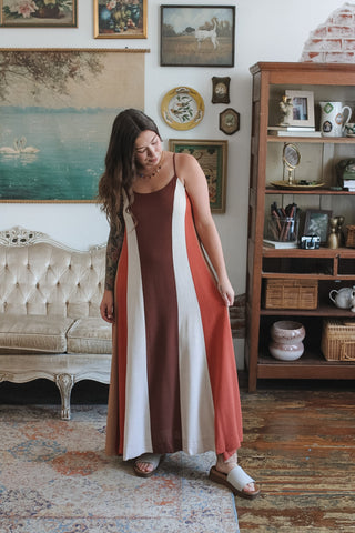 A woman wearing a sleeveless maxi dress with vertical stripes in rust red, cream, and brown, standing in a vintage-style room with a wooden bookshelf and framed art.