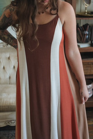 A woman wearing a sleeveless maxi dress with vertical color-blocked stripes in dark brown, white, terracotta orange, and beige, standing in a cozy indoor setting.