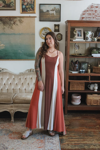 A woman wearing a long sleeveless maxi dress with vertical stripes in terracotta, white, and rust brown, standing in a vintage-style room with a tufted sofa and wooden cabinet.