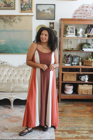 A woman wearing a rust-brown sleeveless maxi dress with vertical white and terracotta side panels, standing in a vintage-style room with a wooden bookshelf and framed art.
