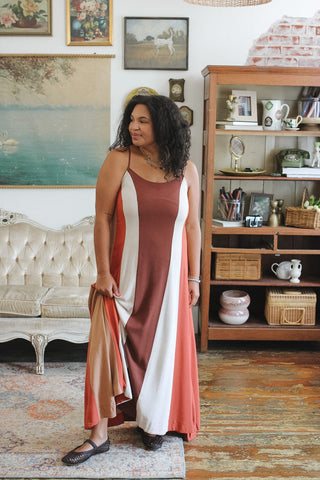 A woman modeling a sleeveless maxi dress with vertical panels of rust brown, white, and terracotta pink in a vintage-style room with a wooden bookshelf and tufted sofa.