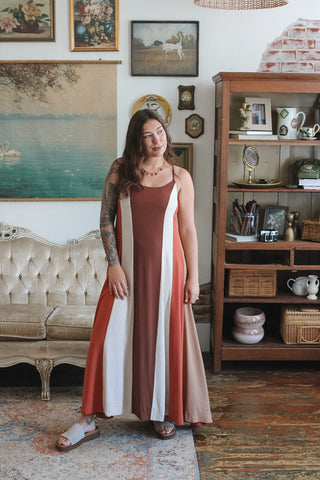 A woman modeling a sleeveless maxi dress with vertical rust brown, white, and beige stripes in a vintage-style room with a tufted sofa and wooden shelving.