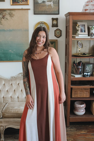 A woman wearing a sleeveless maxi dress with vertical stripes in rust brown, white, and coral pink, standing in a decorated room with a tufted sofa and a wooden bookshelf.