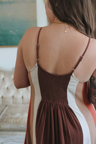 A woman shown from behind wearing a sleeveless smocked dress with thin straps, featuring a deep brown smocked bodice and side panels in dark brown, white, and rust orange.