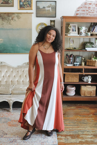 A woman modeling a sleeveless maxi dress with vertical stripes in rust brown, white, and terracotta, standing in a decorated room with a wooden bookshelf and vintage furniture.