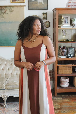 A woman wearing a sleeveless color-block maxi dress with vertical stripes of rust brown, cream, and coral pink, standing in a decorated room with a wooden shelving unit and framed art in the background.