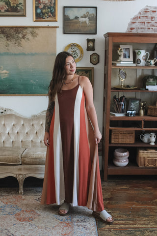 A woman wearing a sleeveless rust-orange maxi dress with white and brown vertical stripe panels, standing in a vintage-style room with a tufted sofa and wooden shelving.