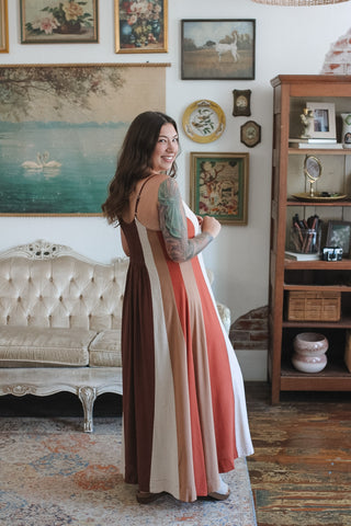 A woman wearing a sleeveless maxi dress with vertical stripes in brown, cream, coral, and white, standing in a vintage-style room with a tufted sofa and wooden shelving.