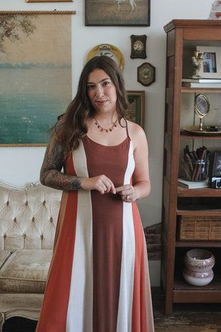 A woman with long brown hair and visible arm tattoos stands indoors wearing a sleeveless maxi dress with vertical rust red, white, and peach panels. The setting is a decorated room with framed wall art, a tufted sofa, and a wooden cabinet.