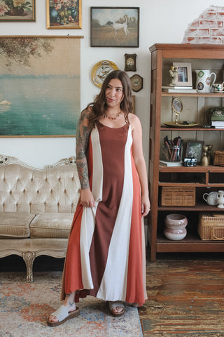 A woman wearing a sleeveless maxi dress with vertical rust brown, white, and terracotta color-blocked panels, standing in a vintage-style room with a tufted sofa and wooden shelving.