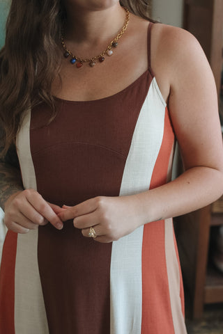 A woman wearing a color-block dress with vertical stripes of dark brown, white, and terracotta orange. She is also wearing a gold chain necklace with small colorful charms and a gold ring.