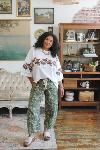 A woman modeling a bohemian-style outfit featuring a white floral embroidered top with red and green floral patterns and green floral print pants, standing in a decorated room with a wooden bookshelf and vintage sofa.