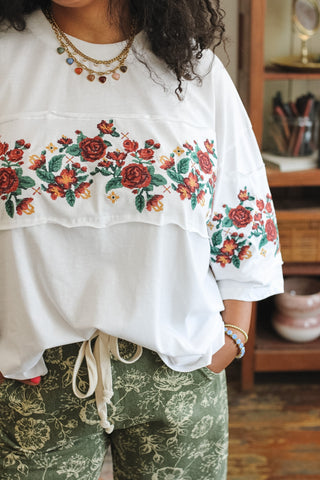 A person wearing a white oversized blouse with colorful floral embroidery across the chest and sleeves, paired with green patterned pants, in an indoor room setting.