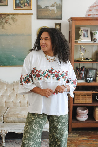 A woman with curly black hair stands in a decorated room wearing a white embroidered top with red and green floral patterns across the chest and sleeves, paired with green patterned pants.