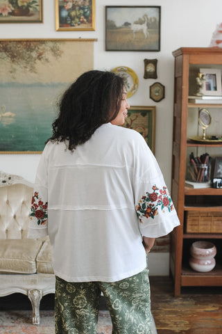 A woman viewed from behind wearing a white short-sleeve tee with red and green floral embroidery on the sleeves, paired with green patterned pants, standing in a vintage-style room with framed art and furniture.