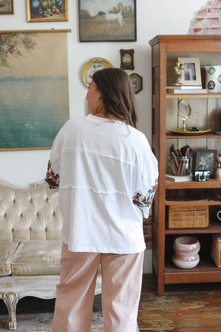 A woman viewed from behind wearing a white tiered tee with colorful floral embroidery on the sleeves and light pink pants, standing in a decorated room with a wooden bookshelf and vintage furniture.