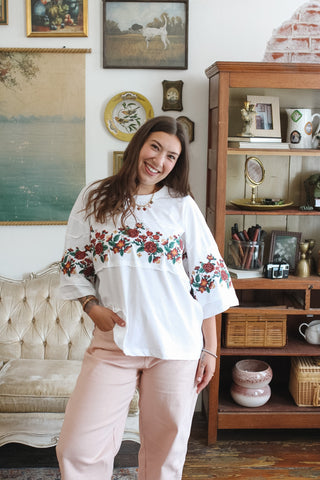 A woman stands in a vintage-style room wearing a white top with red and green floral embroidery across the chest and sleeves, paired with light pink striped pants. She is positioned in front of a wooden display cabinet and a tufted sofa.