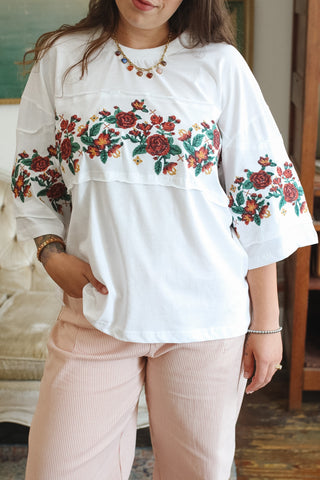 A woman wearing a white short-sleeve top with colorful floral embroidery across the chest and cuffs, paired with light pink striped pants in an indoor setting.