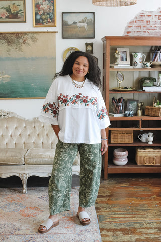 A woman stands in a decorated room wearing a white embroidered top with red and green floral details and green patterned pants.