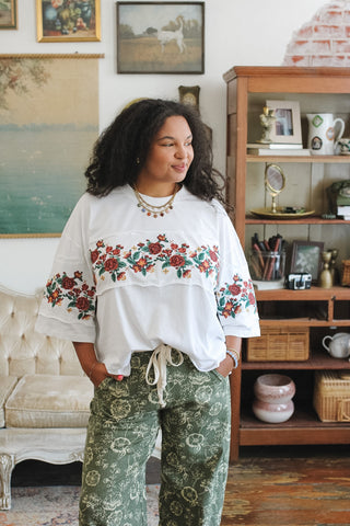 A woman wearing a white embroidered tee with red and green floral patterns on the sleeves and chest, paired with green paisley print pants, standing in a cozy, vintage-filled room.